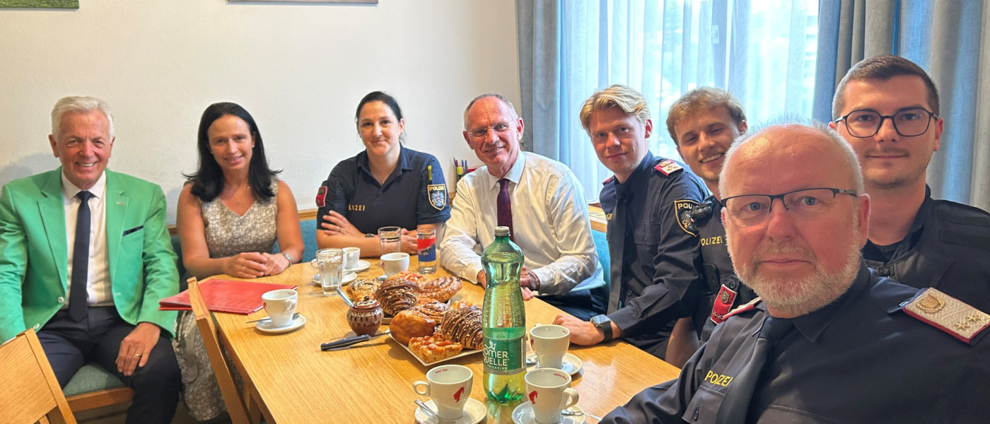 Polizei-Delegation bei Gespräch mit Mann in grünem Sakko und Frau, mit Gebäck und Kaffee.
