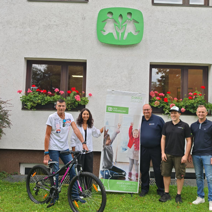 Fünf Personen vor SOS-Kinderdorf-Gebäude mit Spendenbanner und Fahrrad