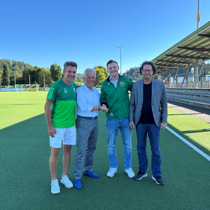 Vier Männer stehen auf einem Fußballplatz, zwei davon schütteln Hände.